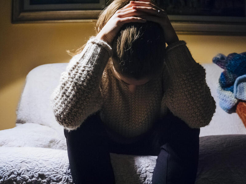 A young woman sitting on the edge of a bed with her hands over her head