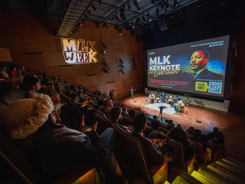 A photo of an auditorium filled with people sitting. At the front of the room are four people sitting in plush chairs in a semi-circle. 