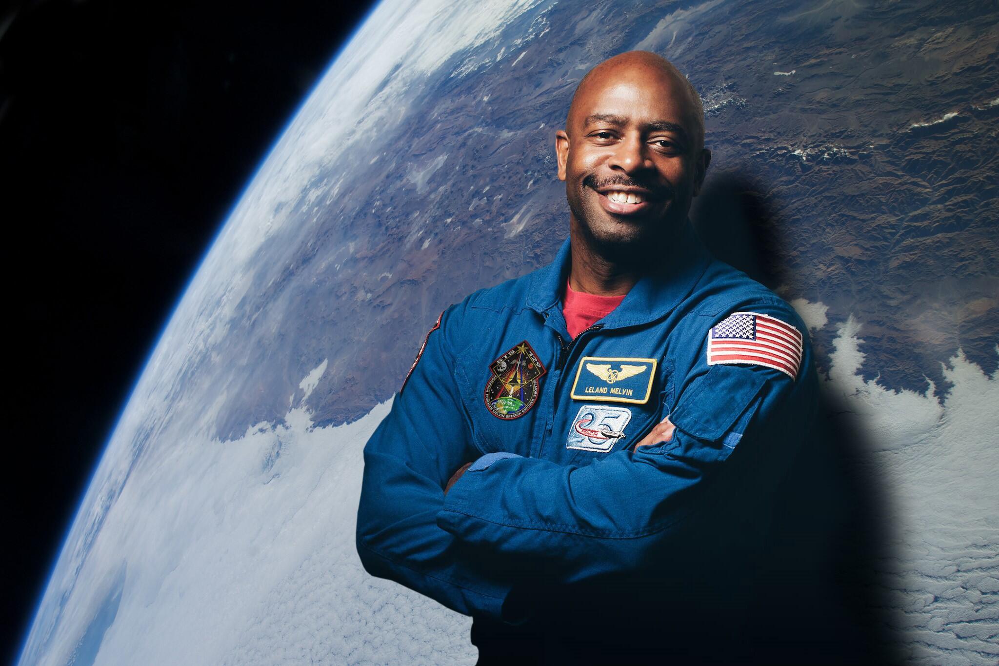 Leland Melvin poses in front of a photo of the Earth taken from space. Patches on his jacket include his nametag, the flag of the United States, the 25th-anniversary patch of the Space Shuttle, and the STS 129 mission patch.
