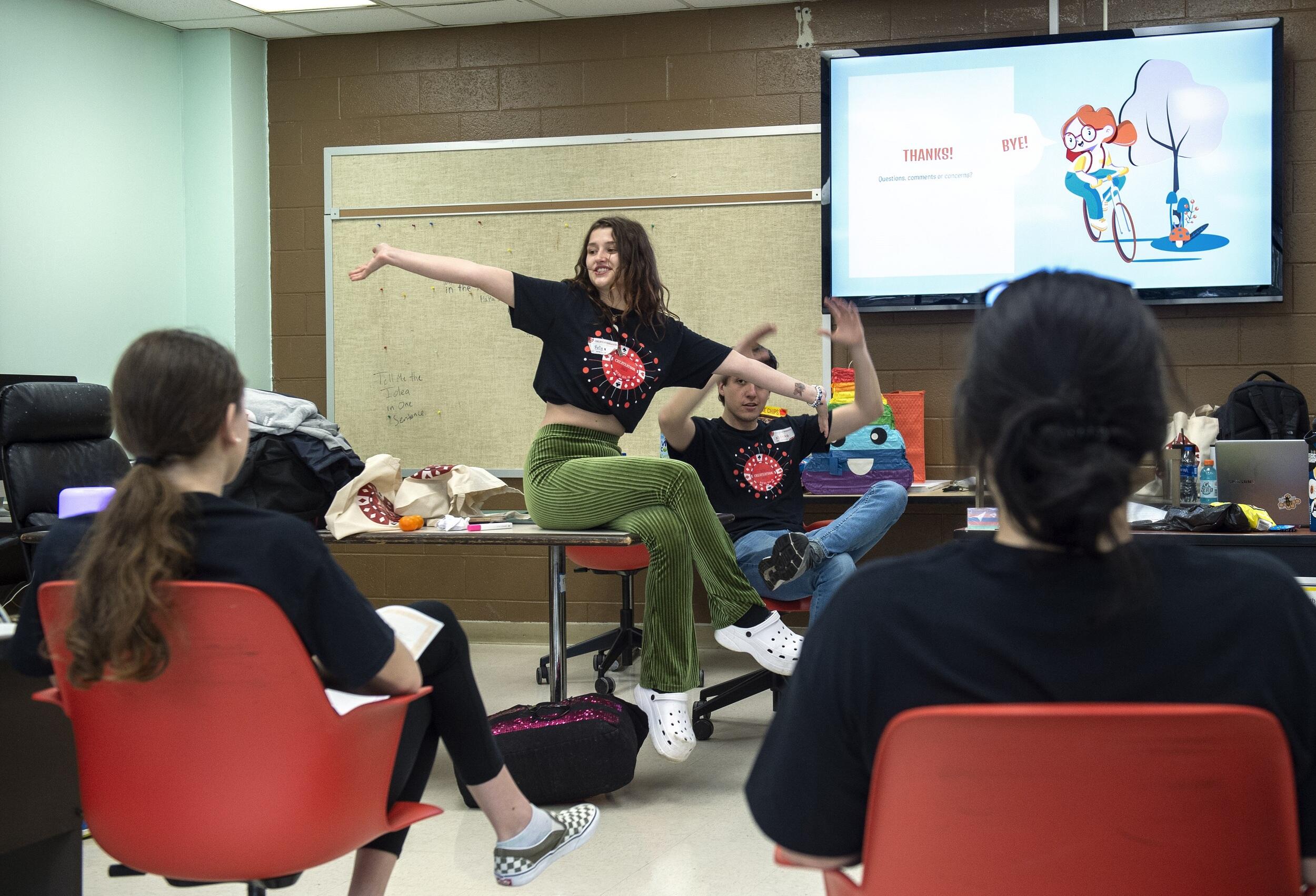 A student sitting on a desk with her arms extended talking to two two others than are seated in front of her. There is another student behind her motioning with his hands in front of a screen 