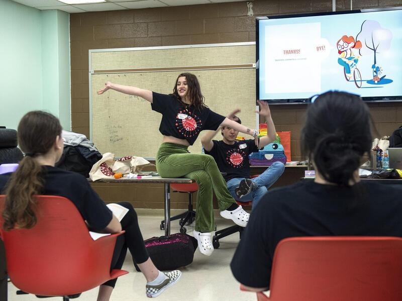 A student sitting on a desk with her arms extended talking to two two others than are seated in front of her. There is another student behind her motioning with his hands in front of a screen 