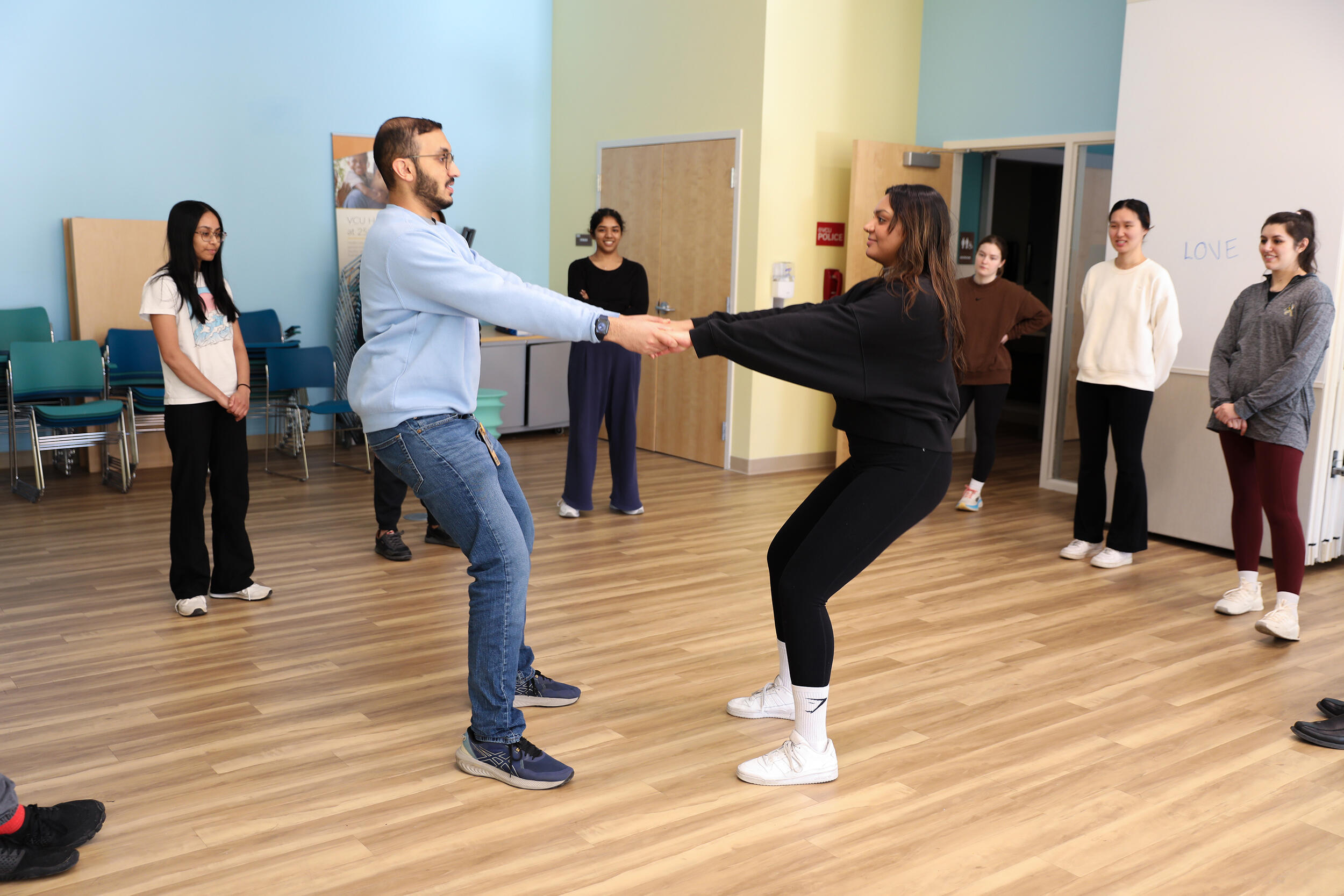 A photo of two people holding hands and leaning their bodies away from each other in the middle of a room. Around the two people is a semi-circle of six other people watching them. 