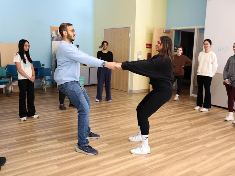 A photo of two people holding hands and leaning their bodies away from each other in the middle of a room. Around the two people is a semi-circle of six other people watching them. 