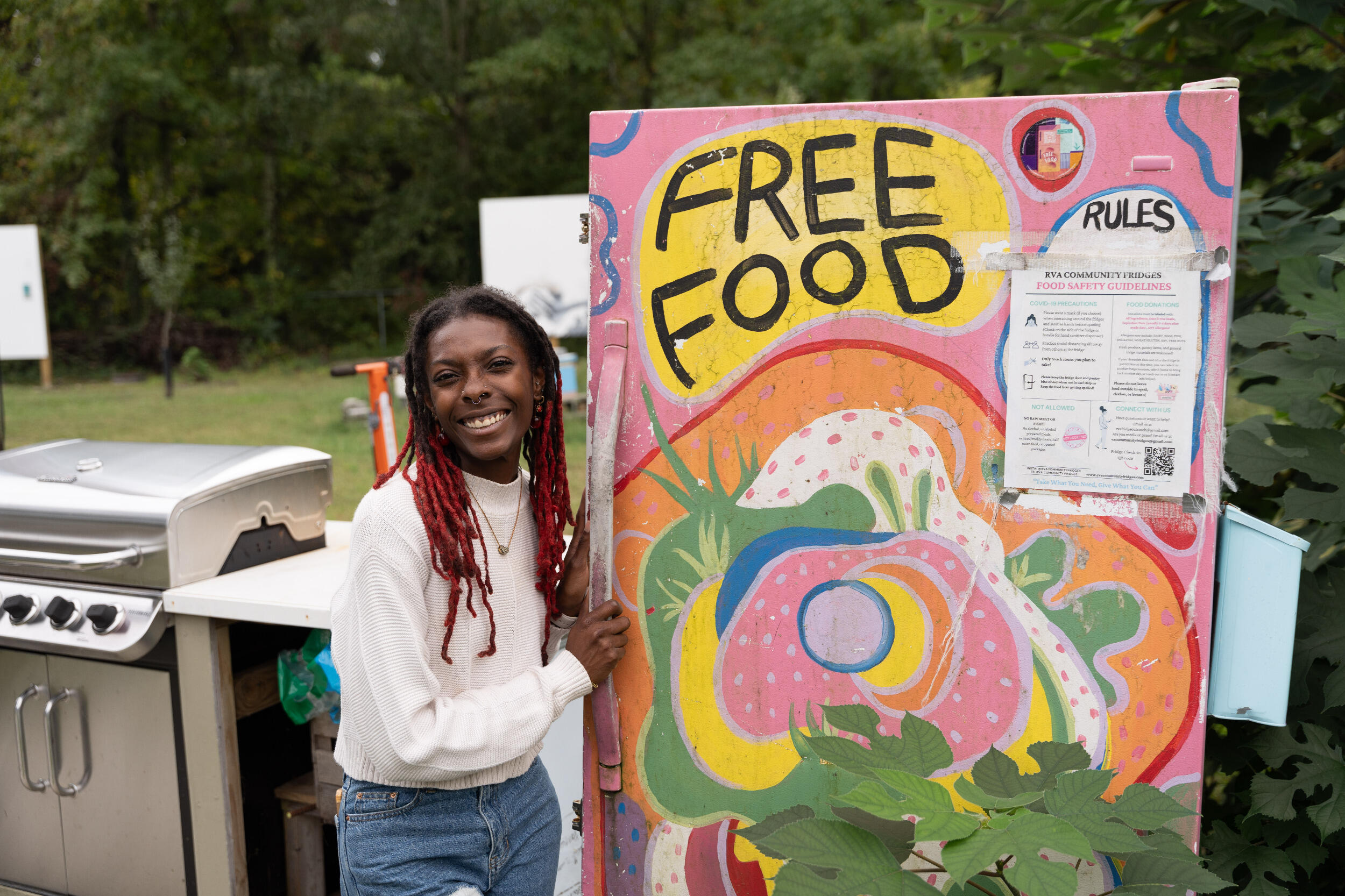A smiling woman stands outside alongside a large sign that says \"Free Food.\"