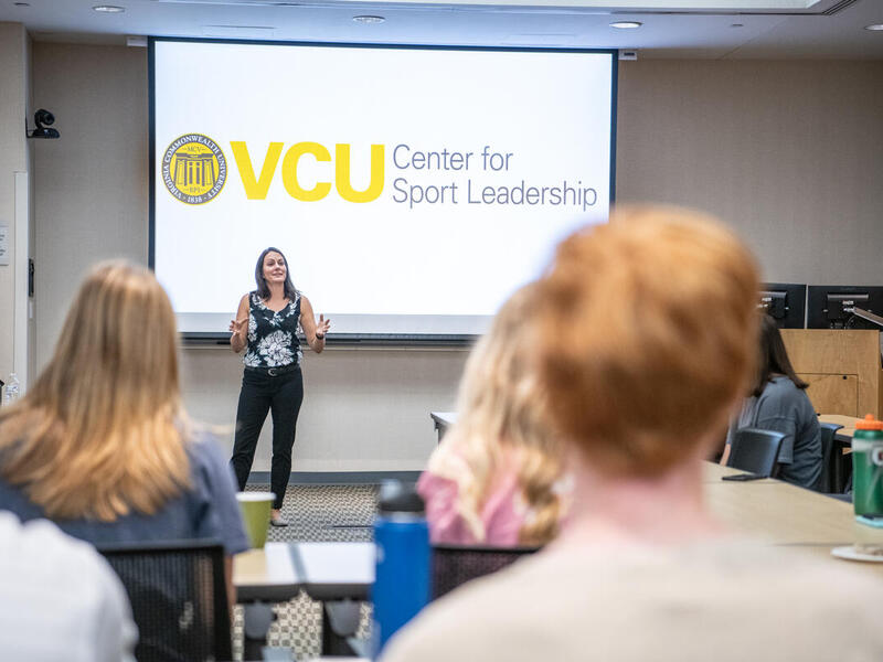 A photo of a woman standing in front of a lecture hall speaking. Behind her is a screen that says \"VCU Center for Sport Leadership\" 