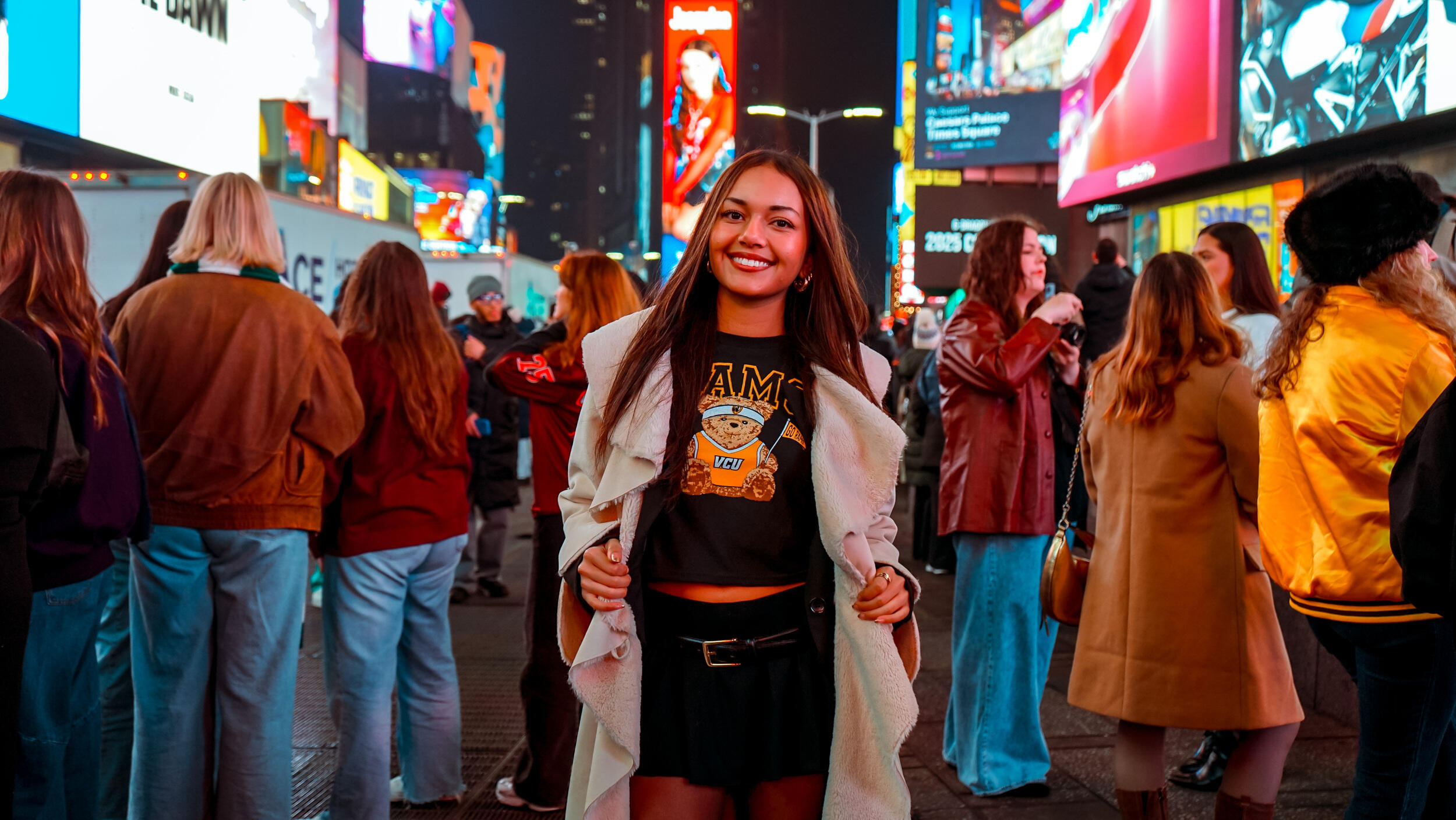 A photo of a woman standing in the middle of Times Square and smiling. She is opening a jacket to reveal a t-shirt that says \"VCU\" and has an illustration of a teddy bear on it. 