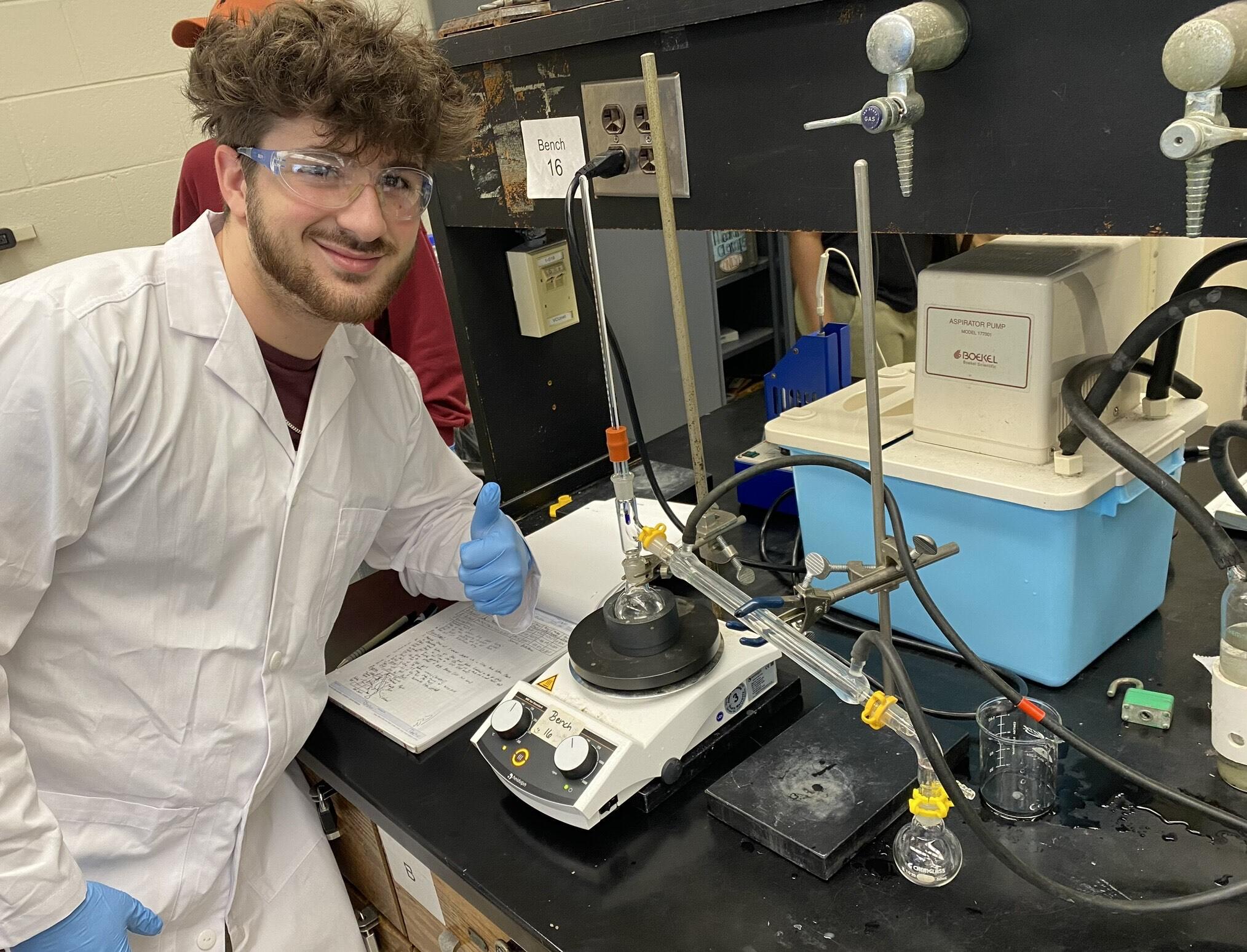 Man in safety goggles and white lab coat leans on counter near research equipment and gives a thumbs up sign.