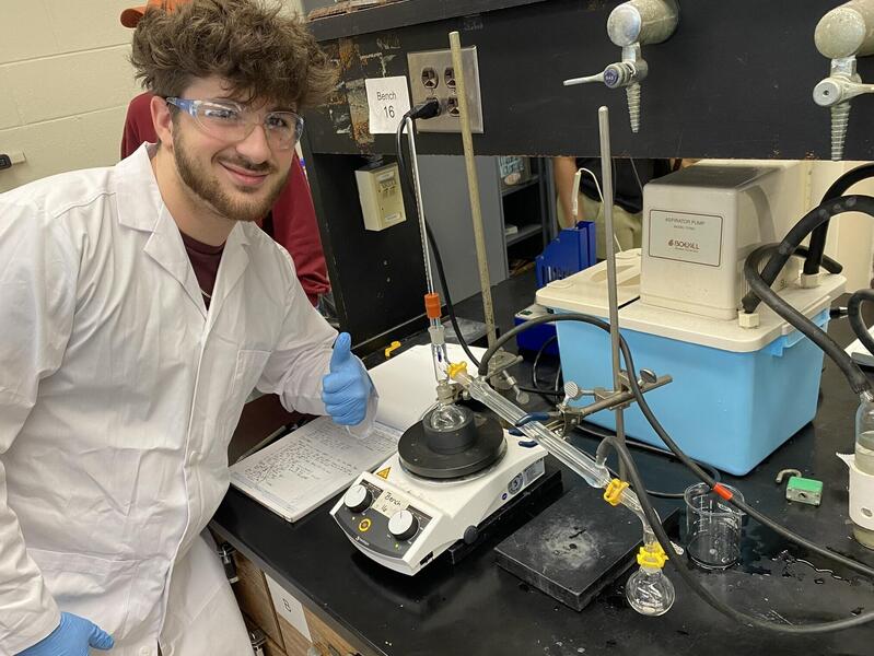 Man in safety goggles and white lab coat leans on counter near research equipment and gives a thumbs up sign.
