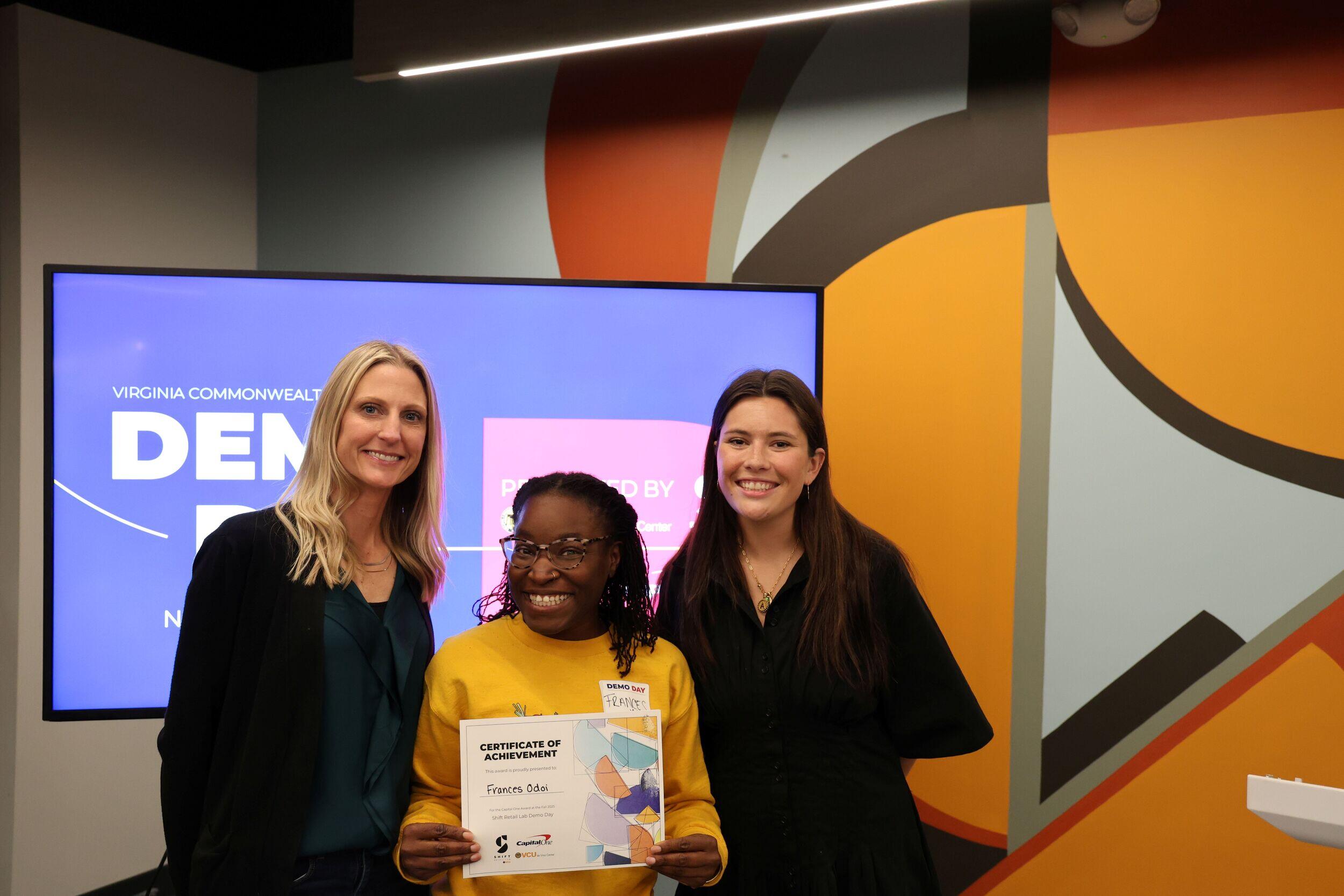 Three women smile in a row. The one in the middle is holding up a certificate.