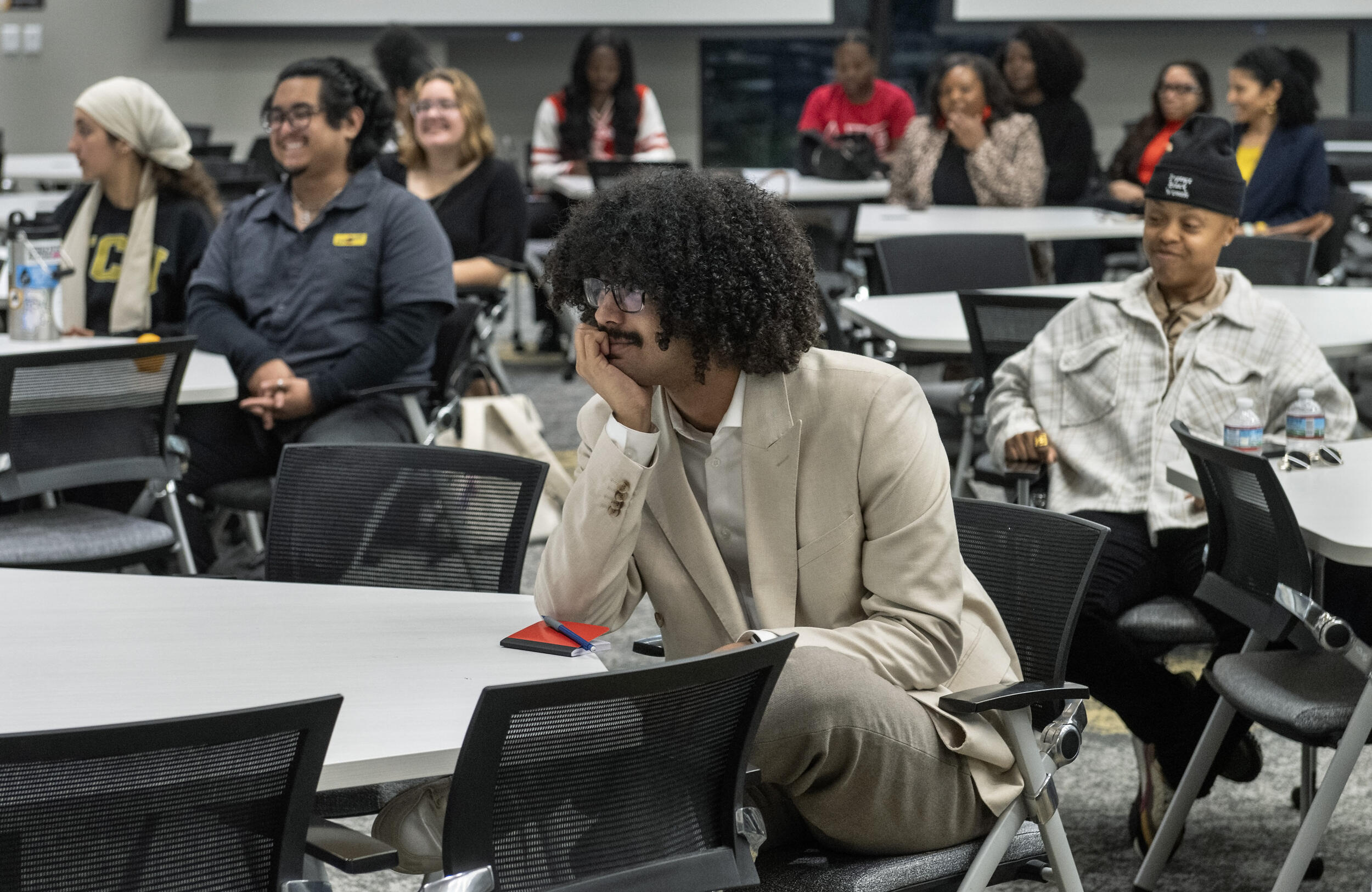 A photo of a room with several circular tables in it. People are sitting in the chairs around the tables and facing left, looking at something in front of them. The man closest to the camera is smiling and resting his chin on his hand. 