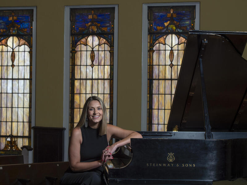 Katie Cappuccio sitting at a piano with stained glass windows behind her