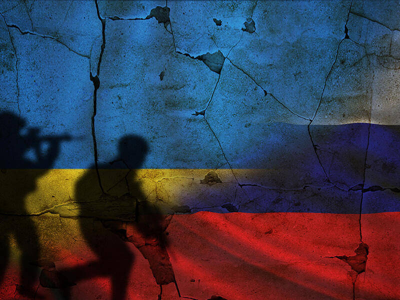 Black silhouettes of two soldiers holding guns in front a cracked brick wall. Half of the wall has the image of the Ukrainian flag and the other half has the Russian flag. 