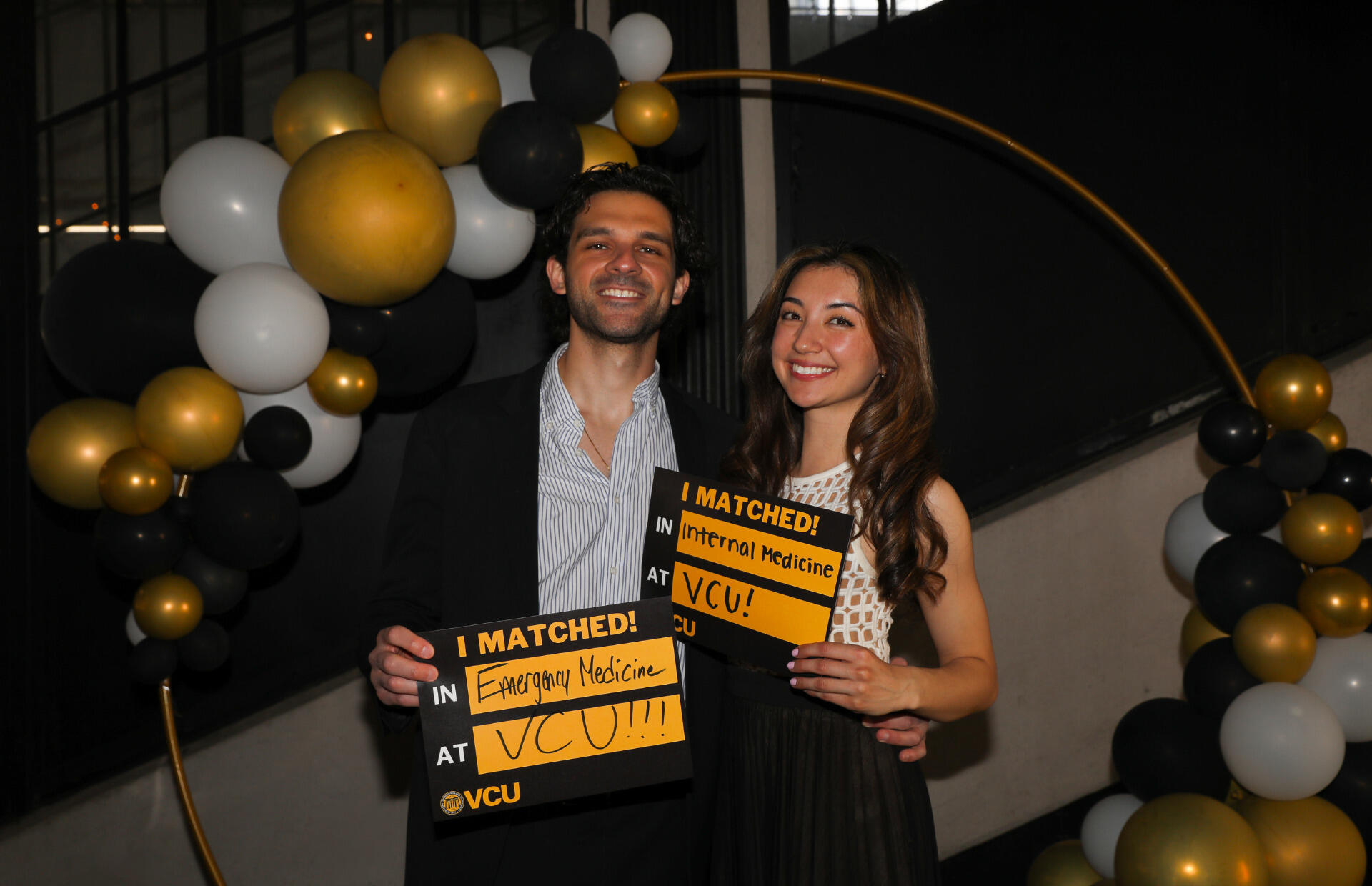 A man and woman standing next to eachother. The man has his right arm around the woman. In the man's left hand is a sign that says \"I MATCHED!\" \"IN Emergency Medicine\" \"AT VCU!!!\" The woman is holding a sign that says \"I MATCHED!\" \"IN Internal Medicine\" \"AT VCU!\" 
