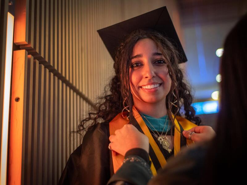 A photo of a woman wearing a graduation cap and gown having another person put a ribbon around her neck. 