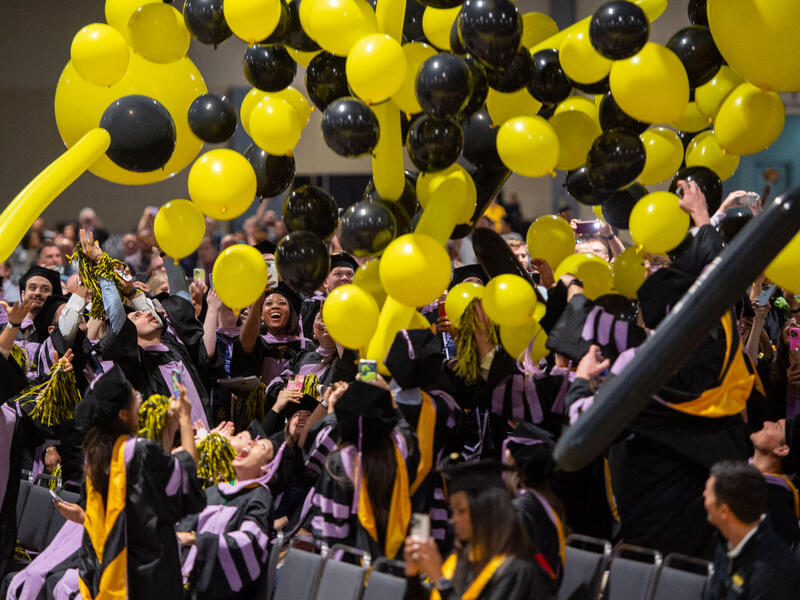 Graduates in cap and gown celebrate beneath yellow and black balloons.