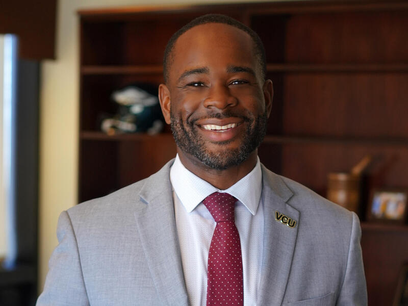 A portrait of a smiling man wearing a light gray sports jacket, white button up shirt, and burgundy tie. On his lapel is a pin that says \"VCU\". 