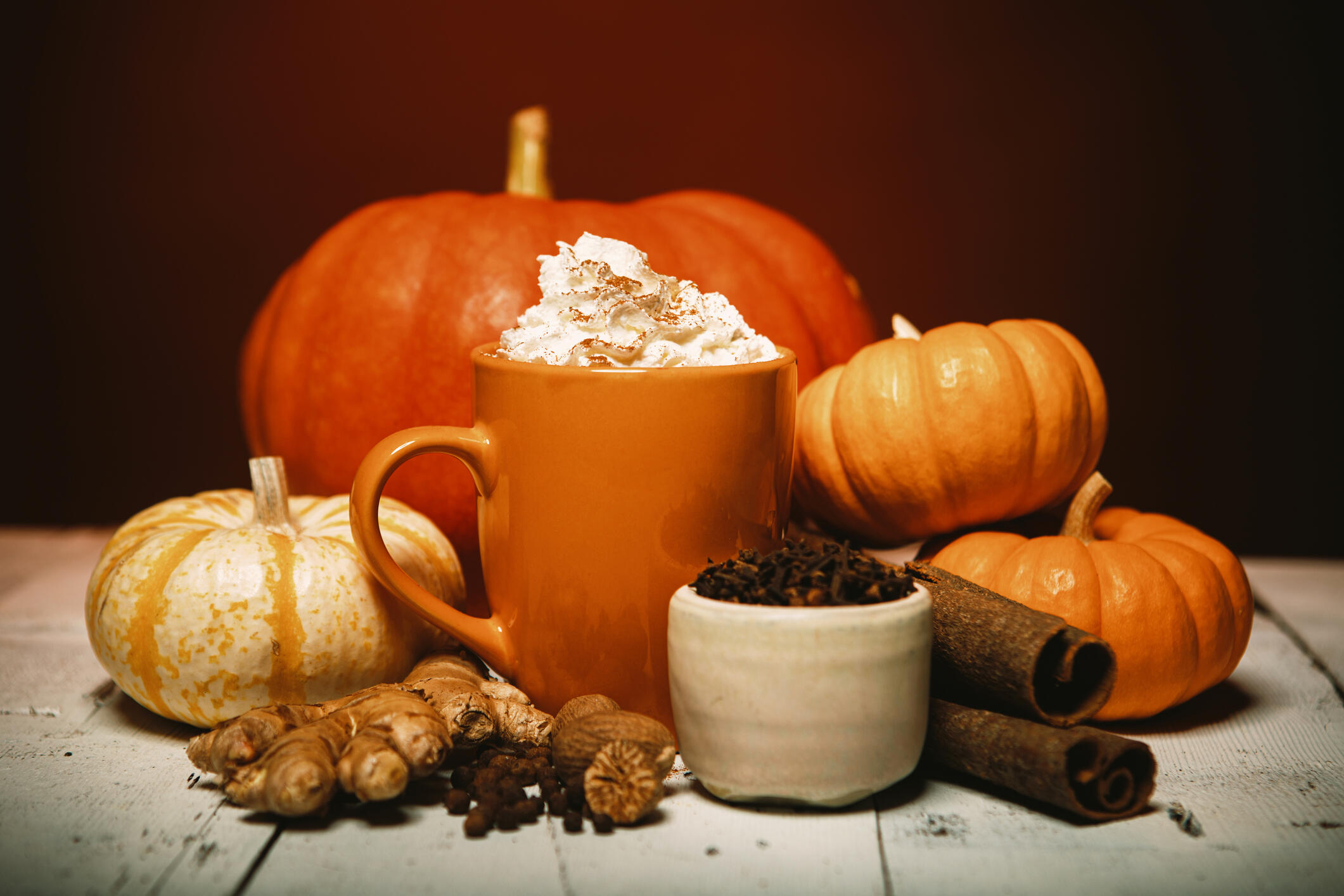 A photo of an orange mug topped with whipped cream surrounded by spices like cinnamon, all spice, and ginger. Behind the cup are four pumpkins. 
