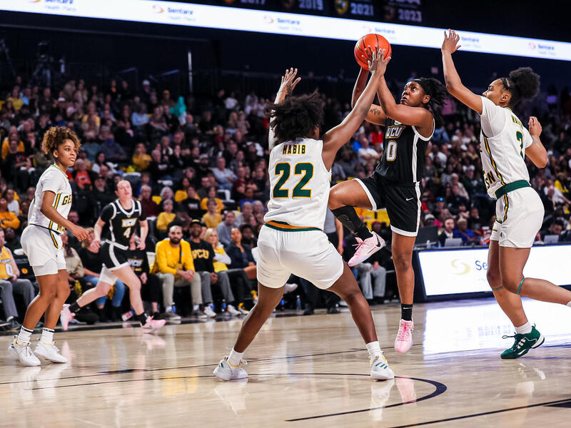 A photo of five women playing basketball on a court. Two women on the right are trying to block another woman from shooting a basket ball. 