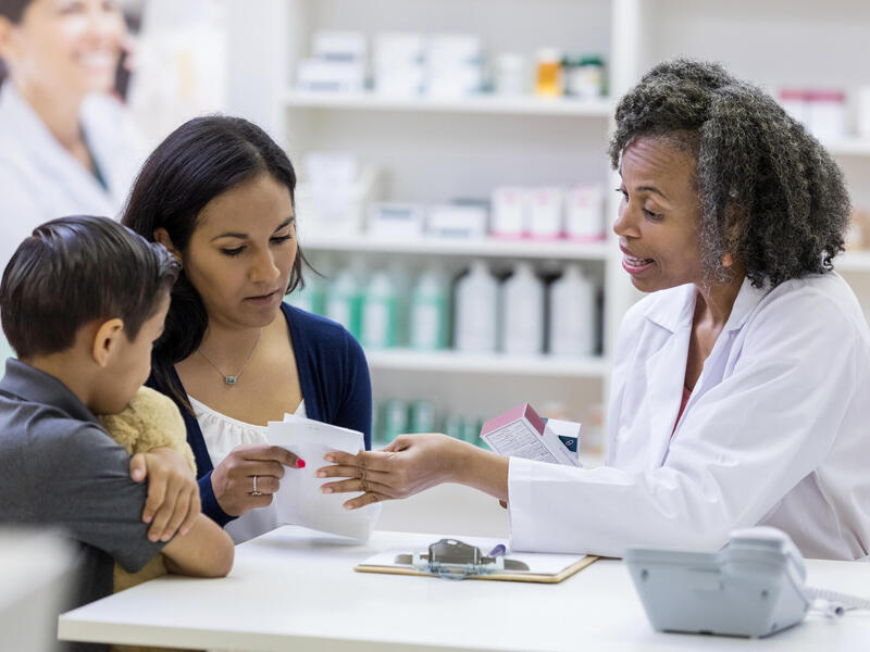 A photo of a pharmacist sitting at a table with a woman and a young boy. The pharmacist and the woman are both holding and looking at the same piece of paper while the pharmacist talks. 