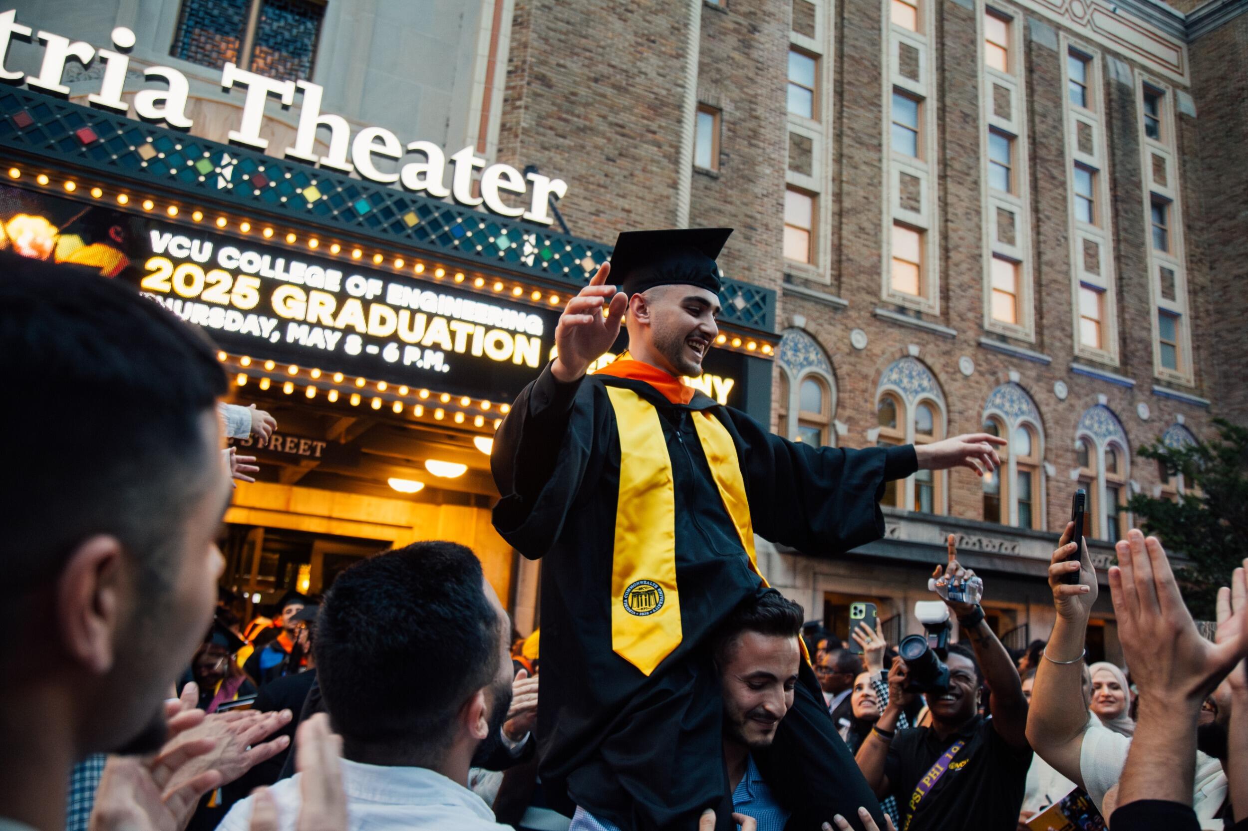 A graduate in cap and gown sits on the shoulders of a man in front of Altria Theater.