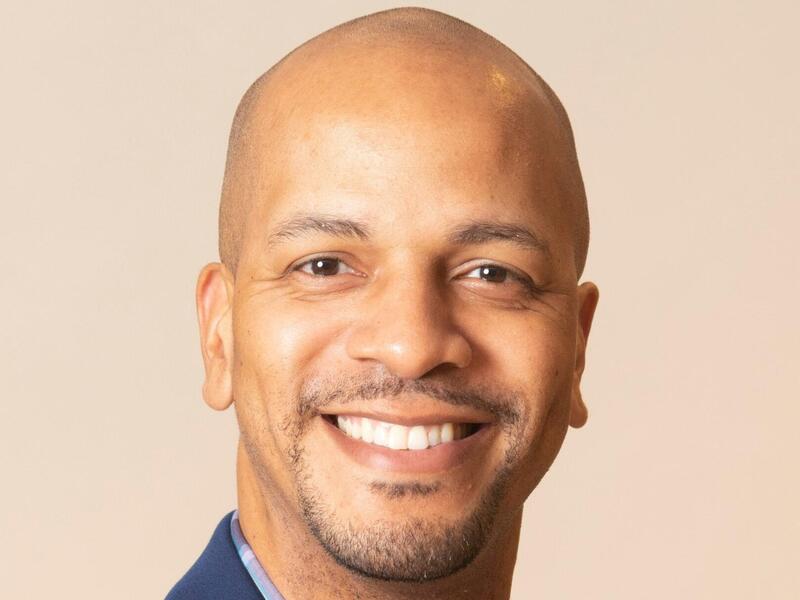 A man smiling wearing a blue sports coat and blue plaid button up shirt. 