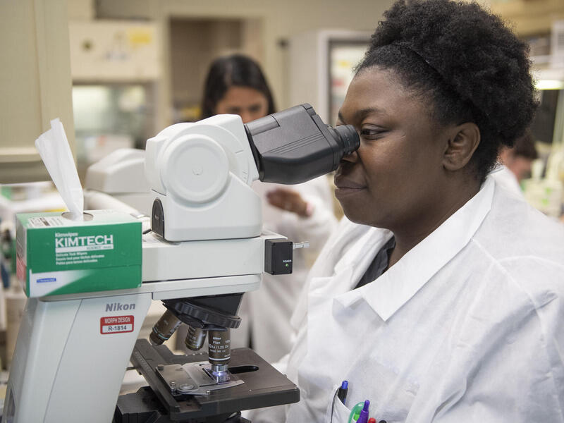 A woman wearing a labcoat looking into a microscope