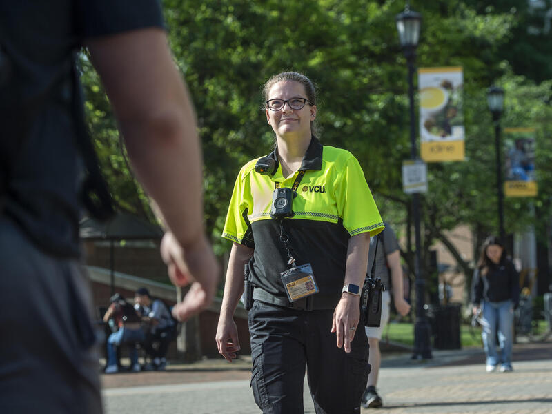 A woman wearing wearing a VCU Police uniform standing in the middle of the sidewalk in a park. 