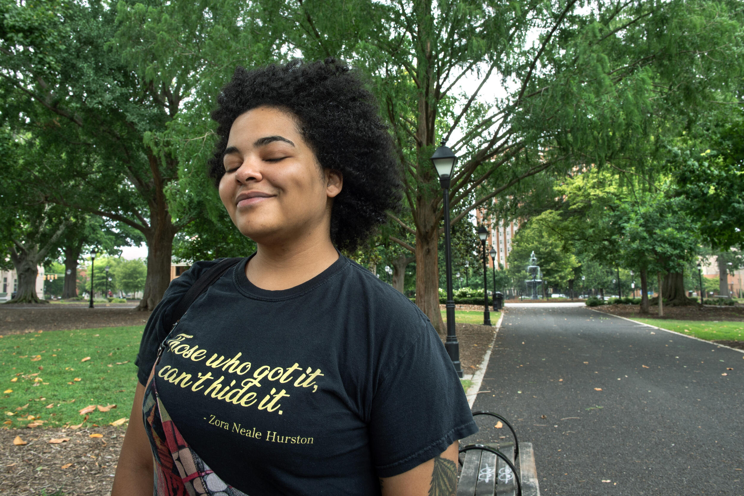 A photo of a person standing on a black path with trees on either side of hit. The person is smiling with her eyes closed. 