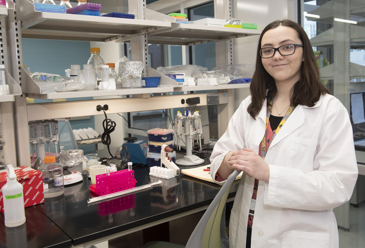 A woman wearing glasses and a white lab coat standing in front of a table with scientific equipment 