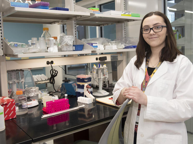 A woman wearing glasses and a white lab coat standing in front of a table with scientific equipment 