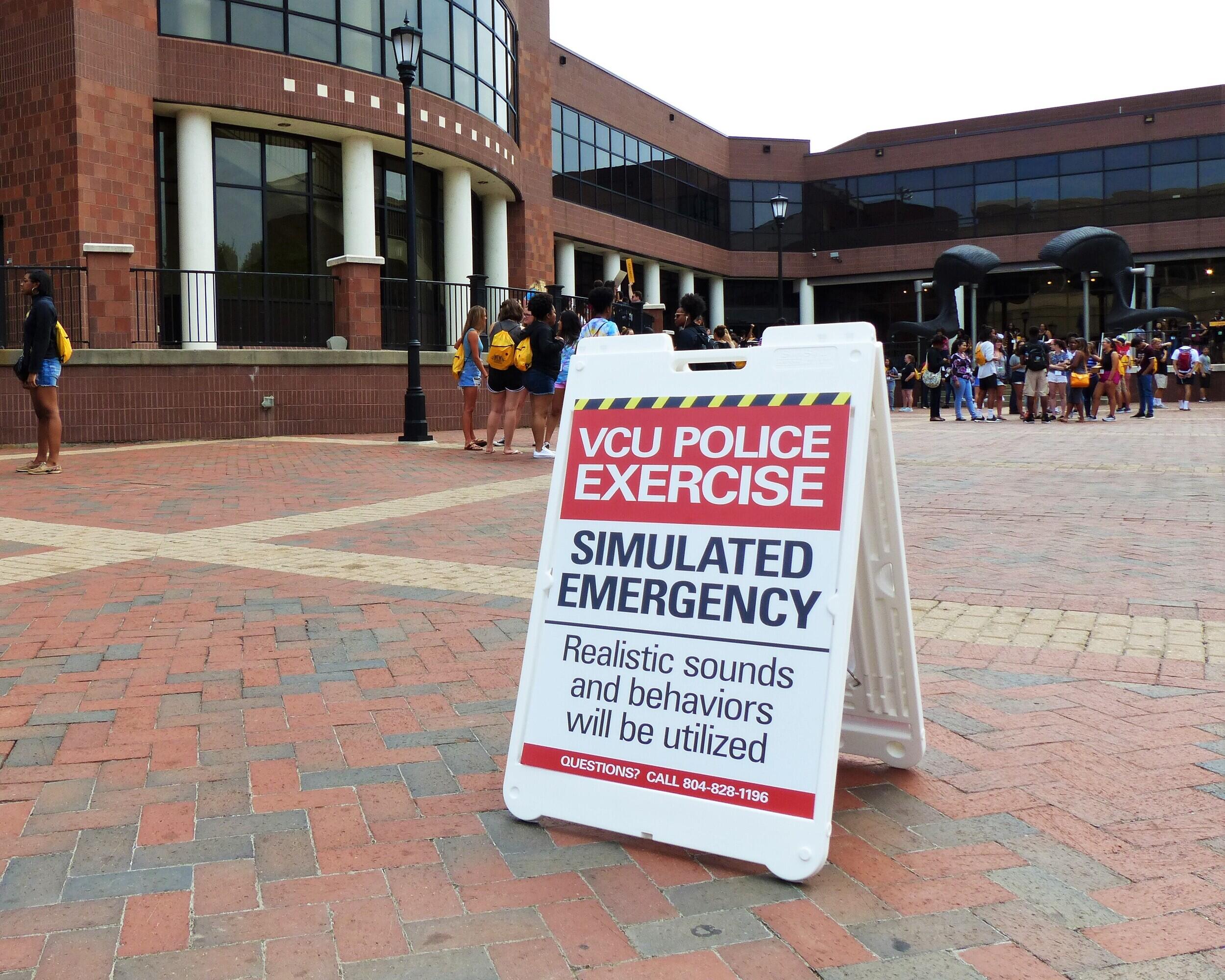 A photo of a sign that says \"VCU POLICE EXERCISE\" \"SIMULATED EMERGENCY\" \"Realistic sounds and behaviors will be utalized>\" Behind the sign is a crowd of people standing around in the distance. 