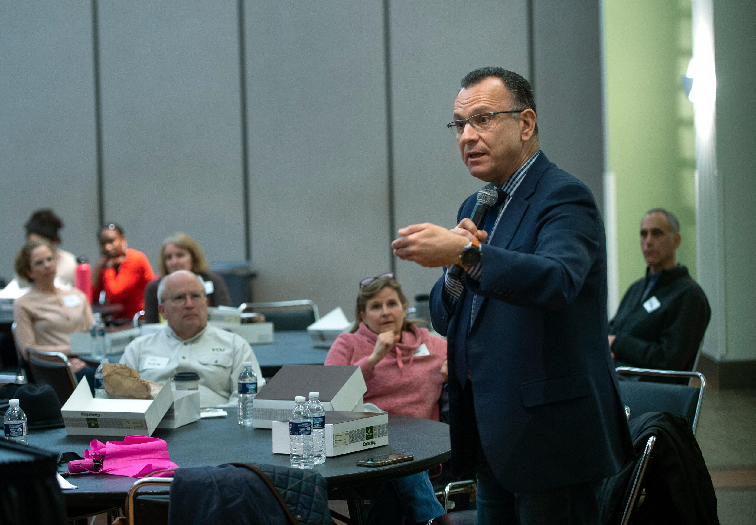 A man stands and gestures while speaking to a group of people sitting at tables.
