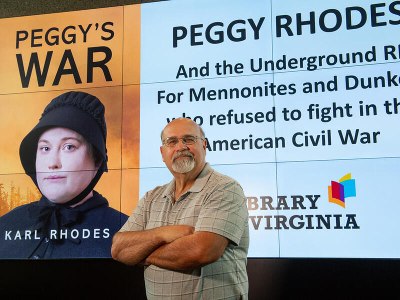 A photo of a man from the waist up standing with his arms crossed against his chest and smirking. Behind him is a screen that says \"PEGGY RHODS And the Underground RR For Mennonities and Dunkers who refused to fight in the American Civil War\" in black text with a photo of the book cover for \"Peggy's War\" next to it. 
