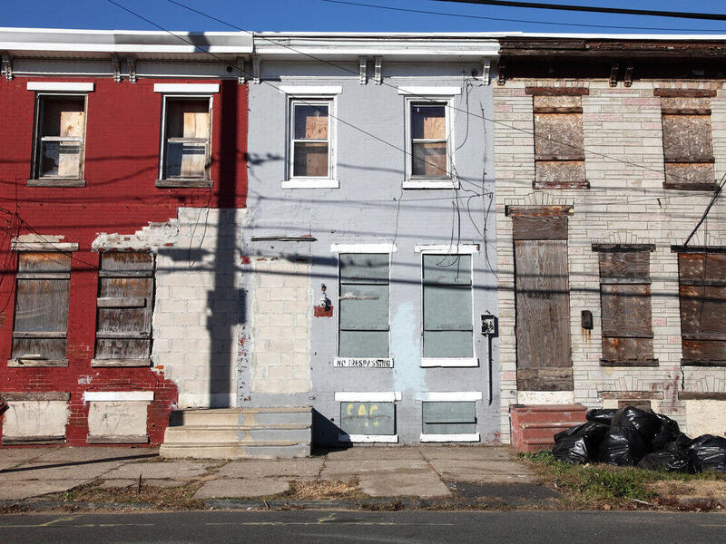 A row of three town houses with the windows and doors boarded up. There's a broken sidewalk and trashbags in front of them. 