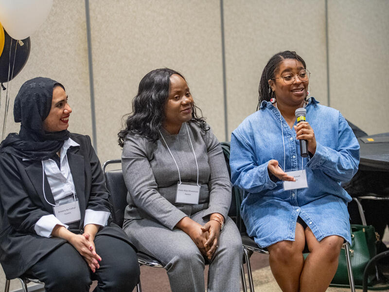 Three women sit side by side. The one on the far right speaks into a microphone.