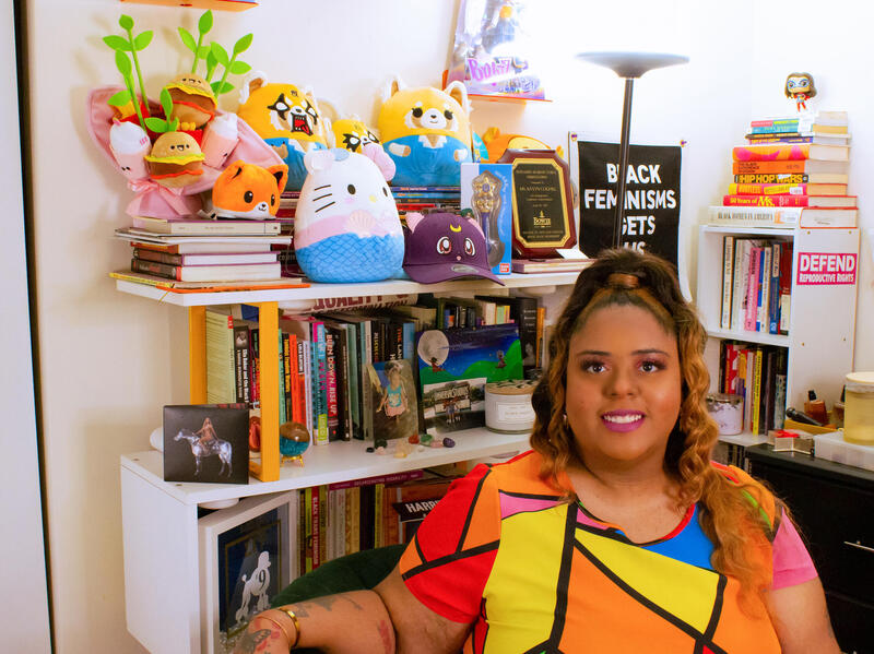 A woman siting in an office filled with books and stuffed toys. 
