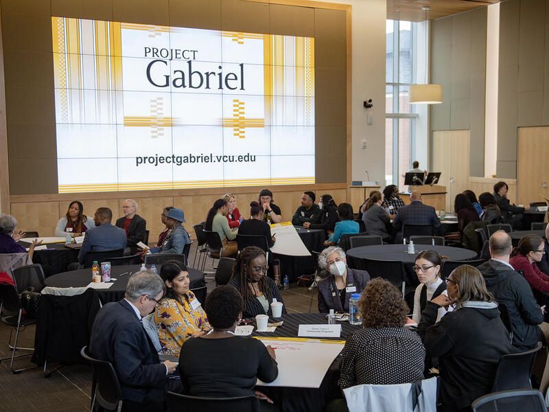 People sitting at circular tables in a large room. There is a screen that says \"PROJECT Gabriel\" behind them. 
