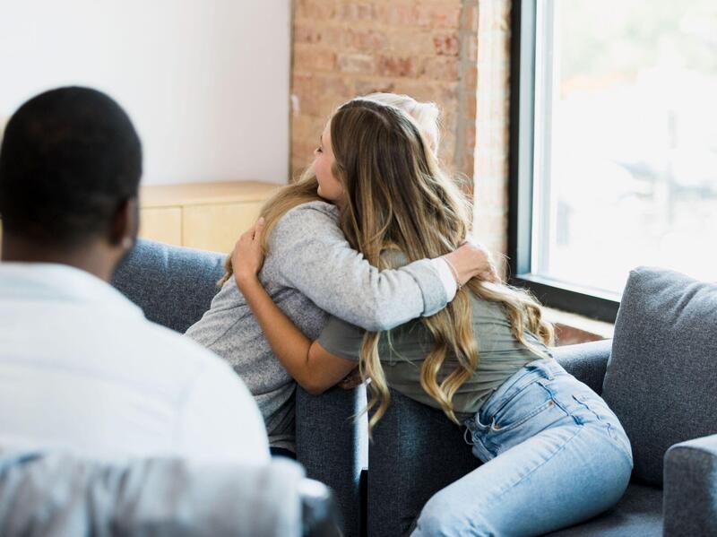 Two people hugging on a couch while a man in a chair sits across from them. 