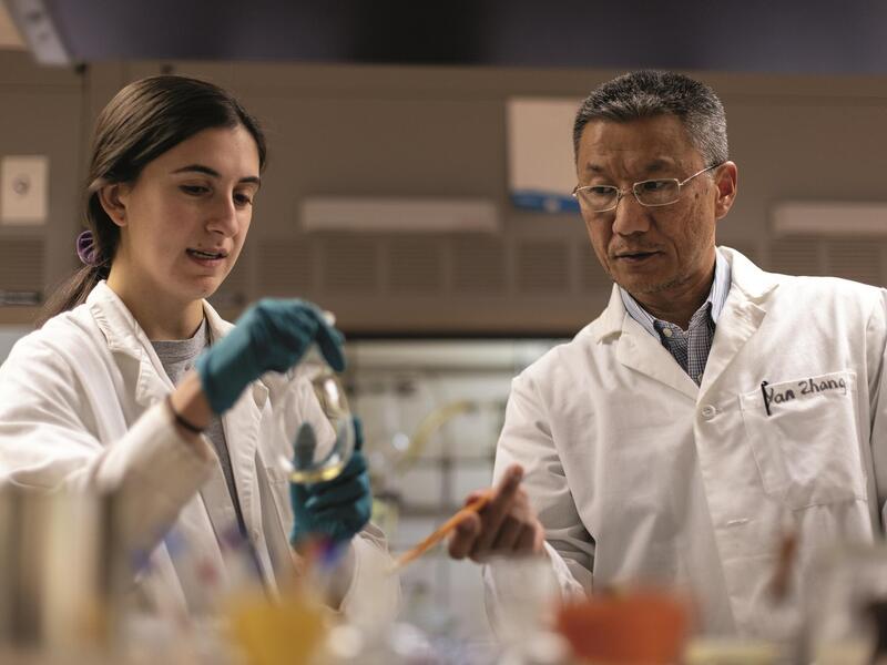 A woman on the left wearing a white lab coat and blue gloves holding a glass object with liquid in it. A man on the right wearing a white lab coat is looking at the woman