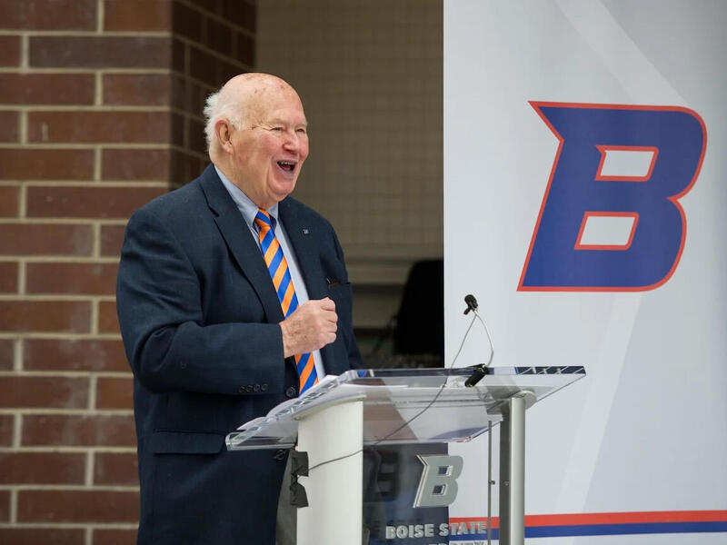 A photo of a man from the waist up. He is standing behind a podium with a microphone on it. Next to him is a white banner with the letter \"B\" on it. The letter is blue with a think orange outline. 