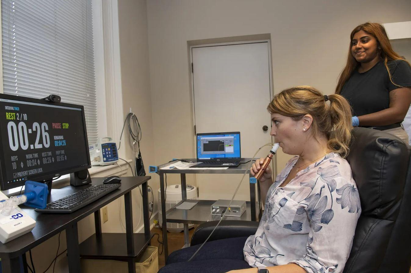 A woman breathing into a tube in front of a screen with numbers on it. Another woman is standing next to her. 