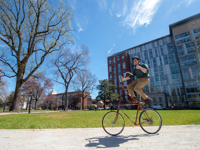 Man on tall bike riding through Monroe Park while giving the thumbs up.