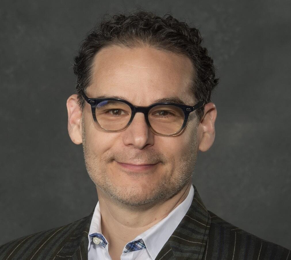 A photo of a man wearing a pin striped suit jacket, a light blue button up shirt, and black glasses. 