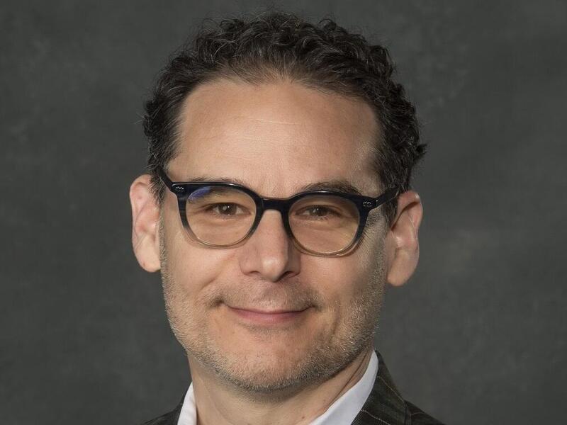 A photo of a man wearing a pin striped suit jacket, a light blue button up shirt, and black glasses. 