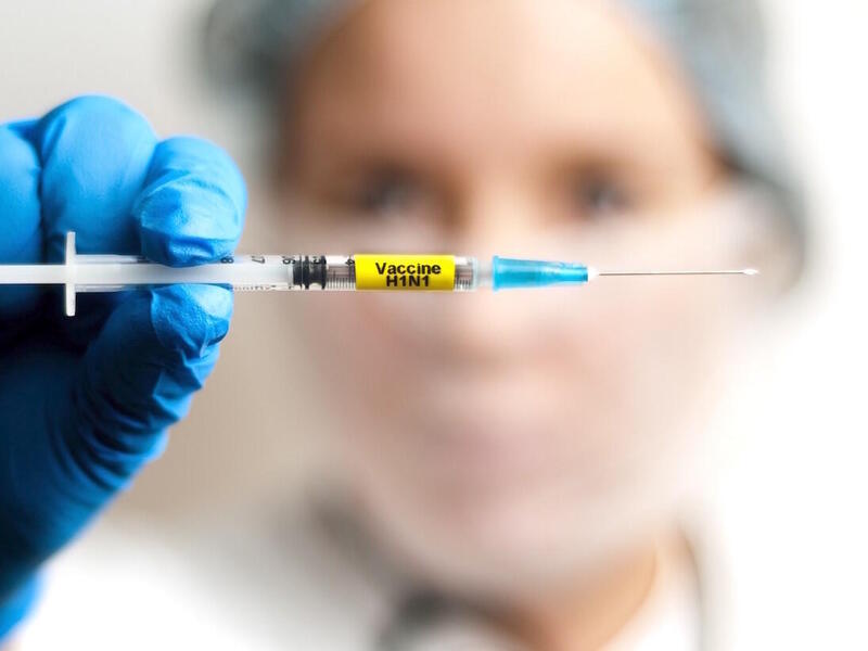 Person in medical protective gear holding a syringe labeled H1N1.