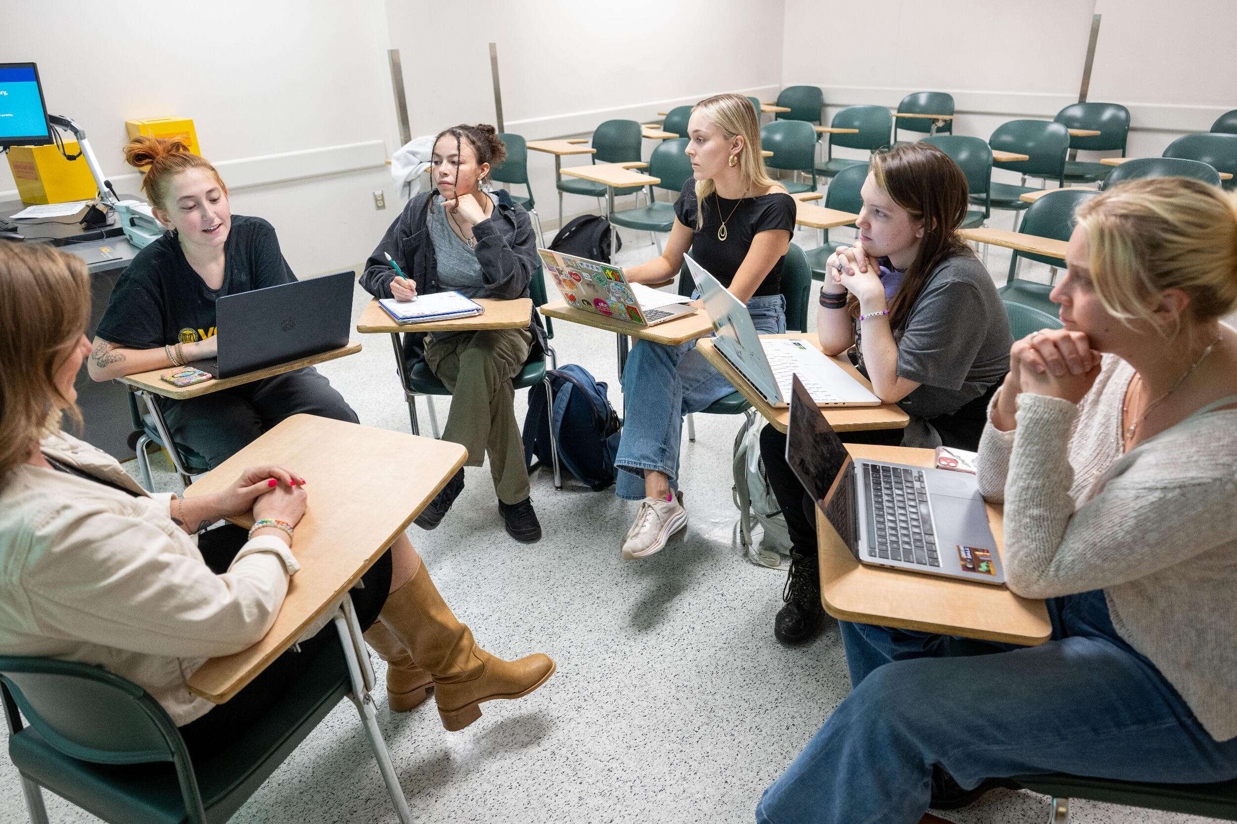 Six people sit at desks in a circle in a classroom and talk.
