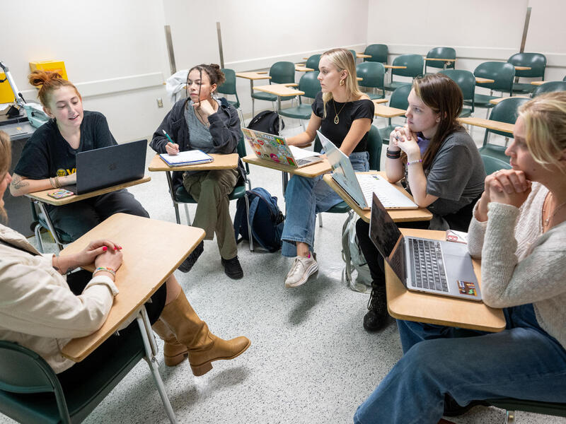Six people sit at desks in a circle in a classroom and talk.