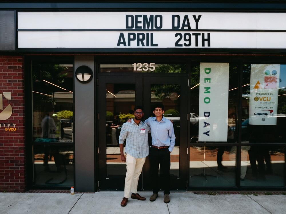 (Left to right) Pranav Neyveli and Miguel Rao standing with one arm around each other under a sign that says \"Demo Day April 29th.\" 