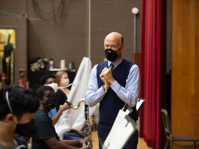 a symphony conductor, speaks to his musicians during a rehearsal. everyone is wearing a mask