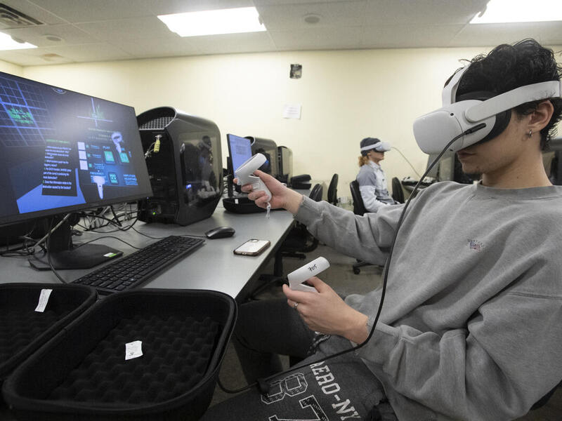 A photo of a person sitting at a desk with a computer monitor on it. The person is wearing a VR headset. 